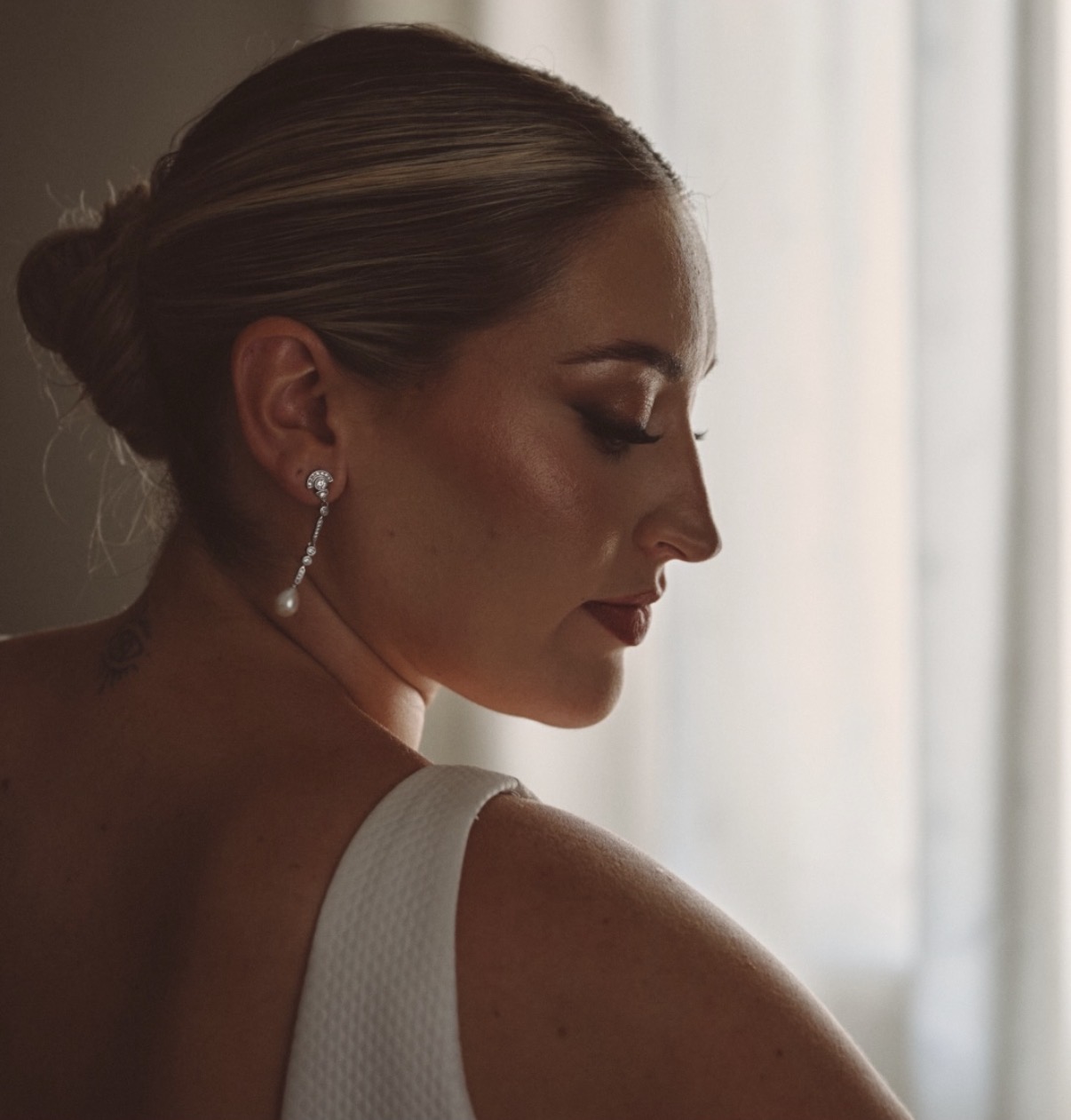 Screenshot Mujer con maquillaje elegante, peinado recogido y pendientes largos, vista de perfil con luz suave.
