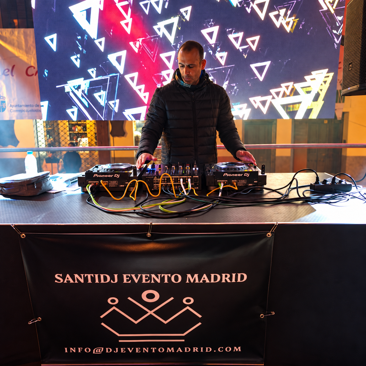 95f4ed18-363a-46e3-9c9e-95c9f3e0d308 Dj con chaqueta negra mezclando música en consola frente a pantalla con luces y gráficos geométricos.