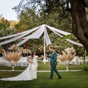 Novios caminando bajo un dosel blanco en un jardín con sillas y decoración natural para boda.