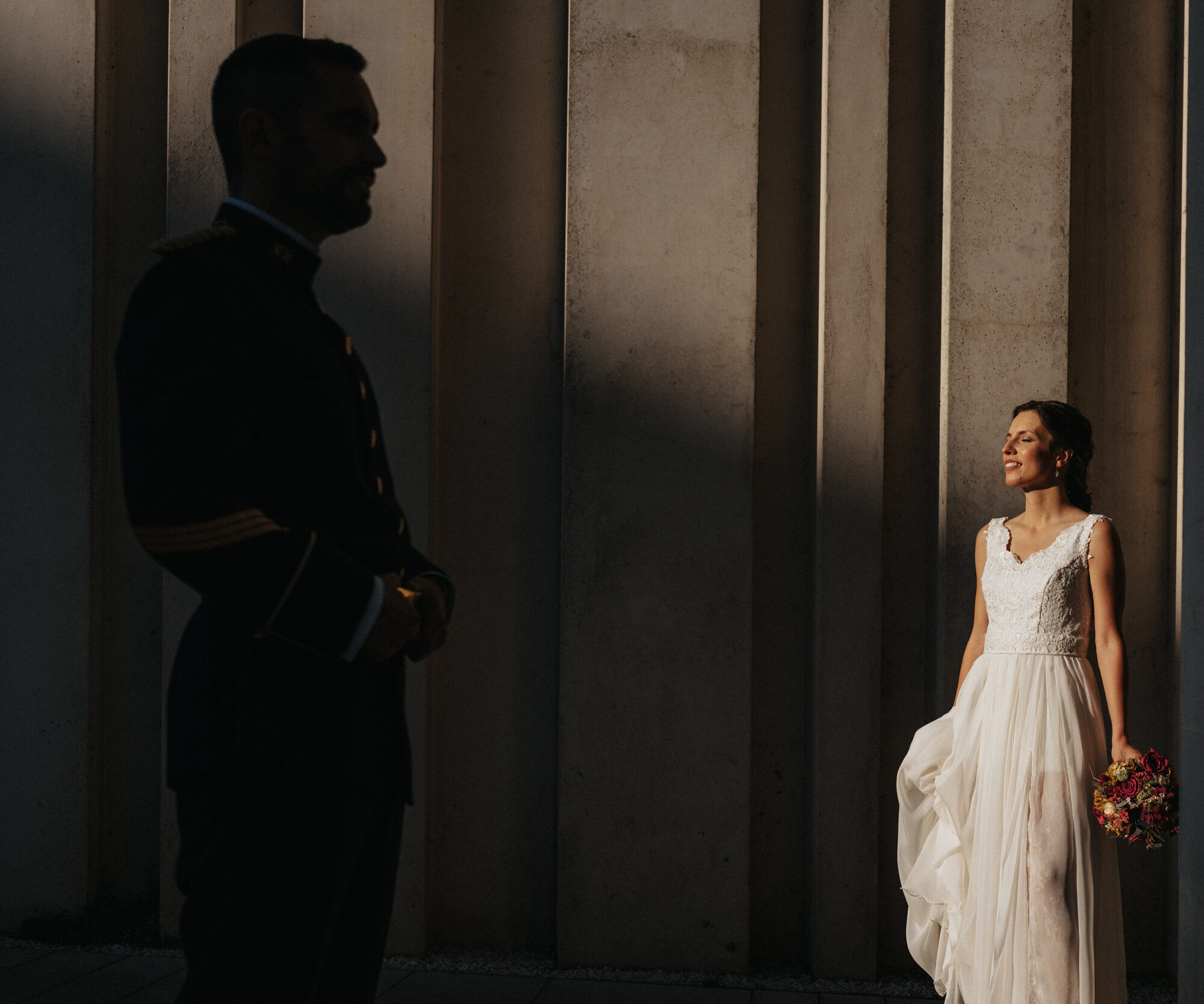_JOF4273 Novia con vestido blanco y ramo iluminada, novio en sombra con traje oscuro frente a columnas.