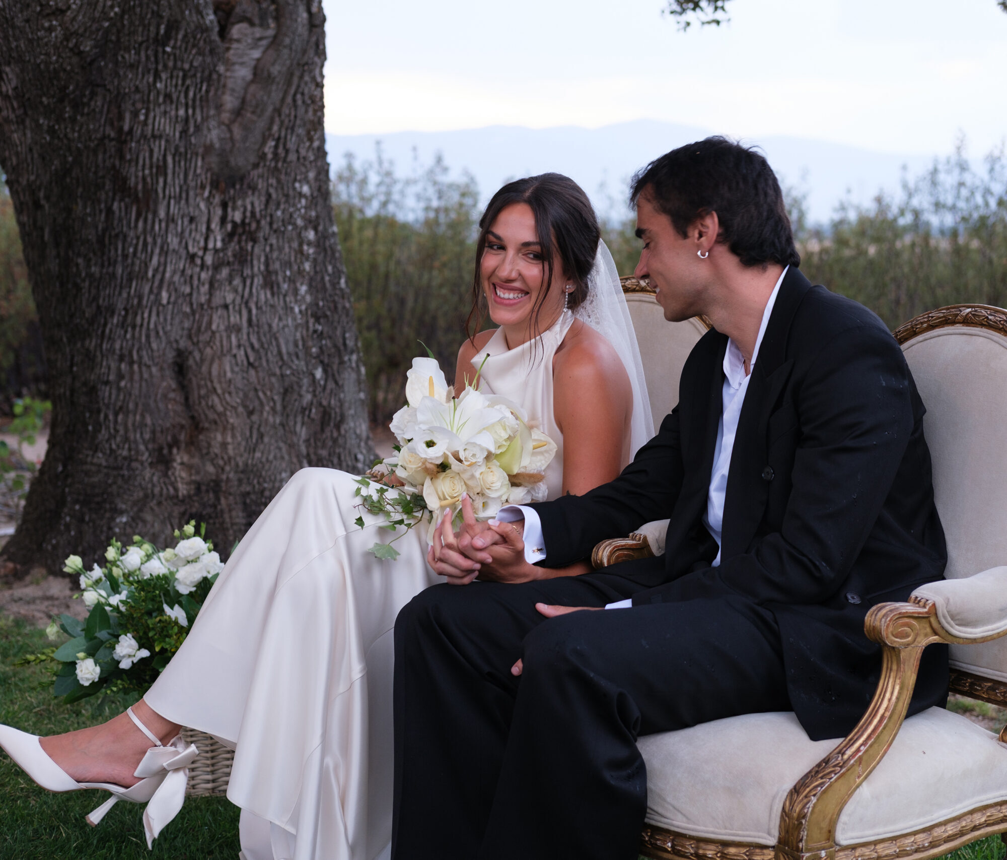 J_y_C_135 Pareja sonriente sentada en sillones antiguos al aire libre, ella con vestido blanco y ramo de flores.