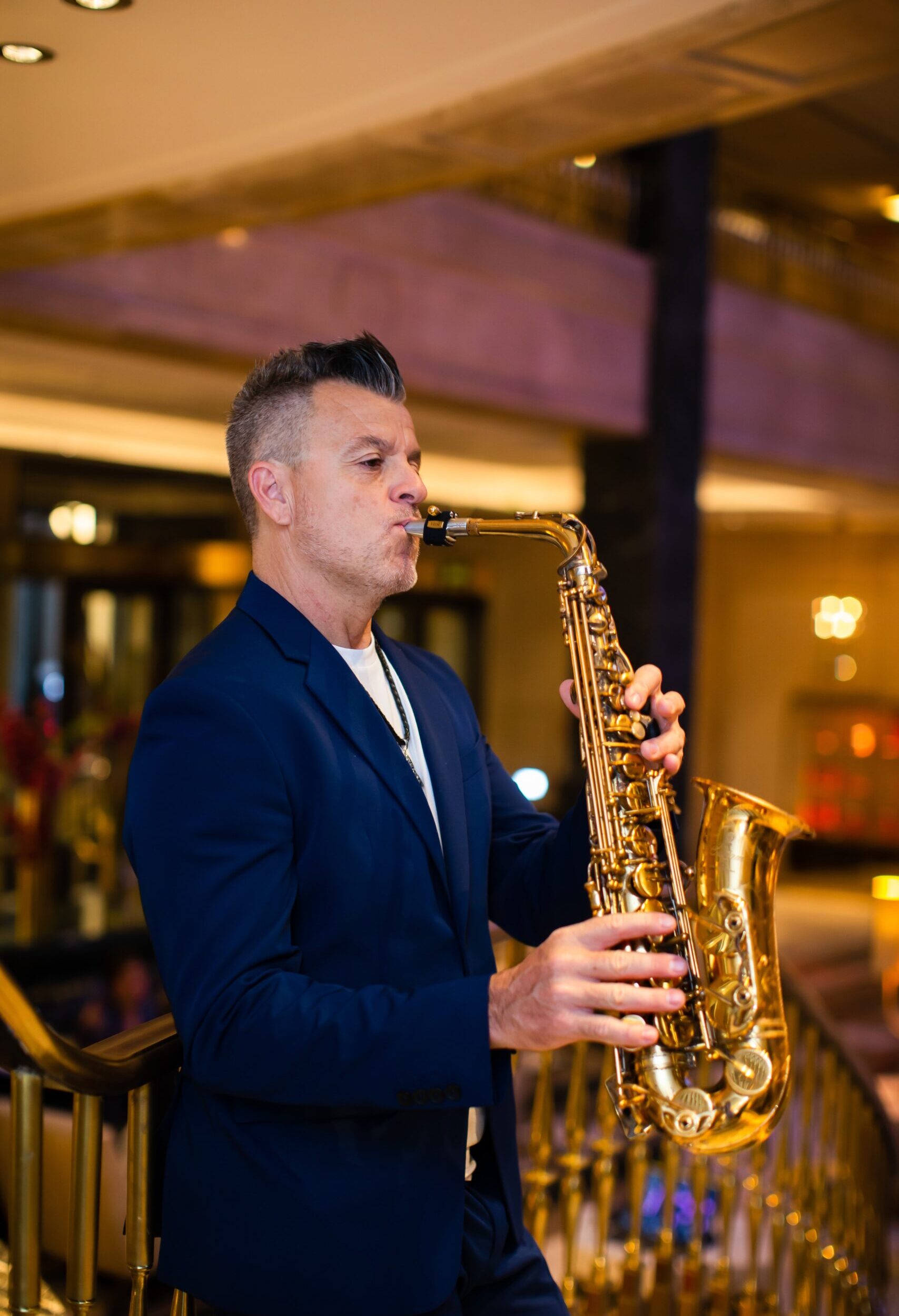 NICOLETA_RAULSAXO-36 Hombre tocando saxofón en un espacio interior con luces cálidas y barandilla dorada.