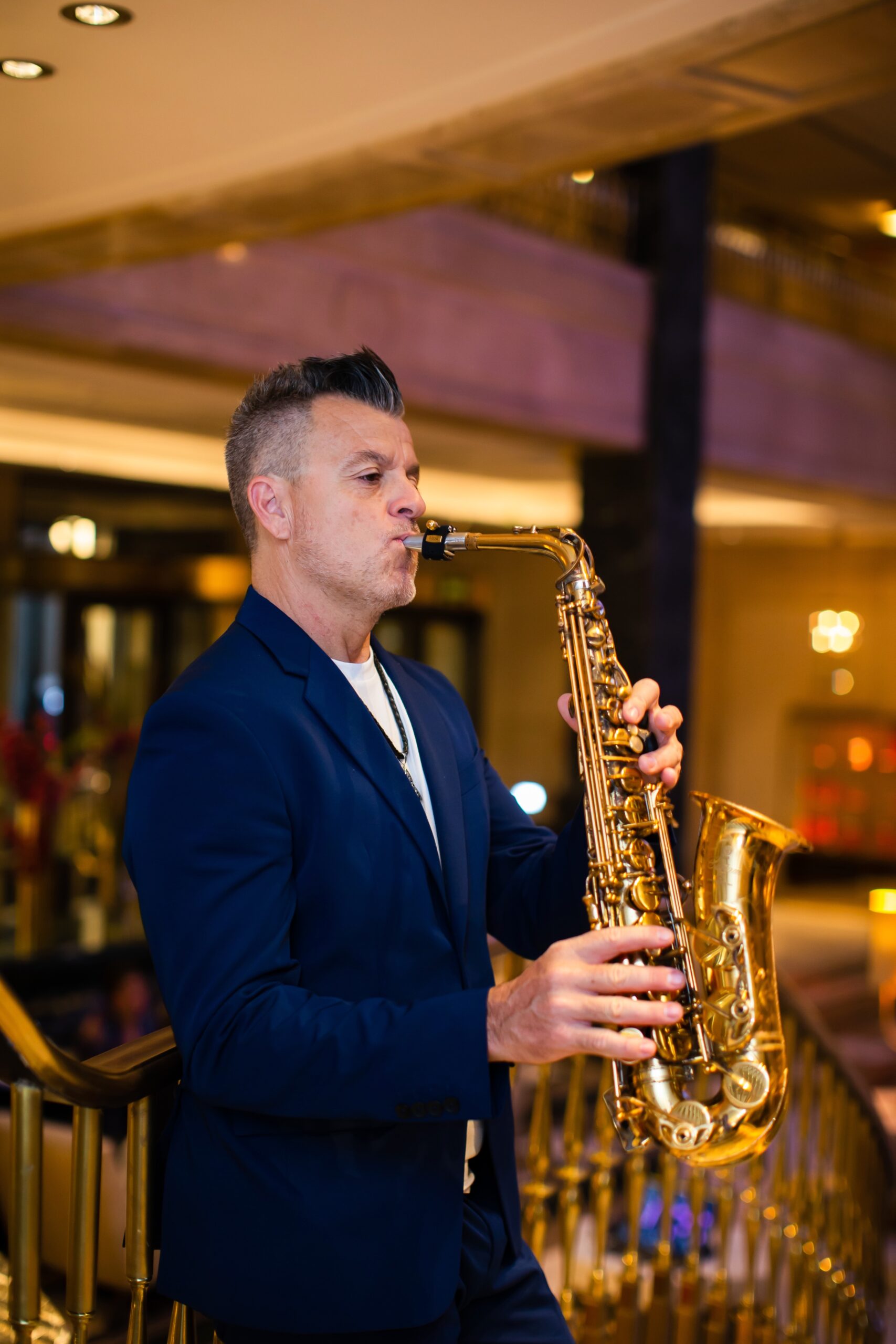 Hombre tocando saxofón en un espacio interior con luces cálidas y barandilla dorada.