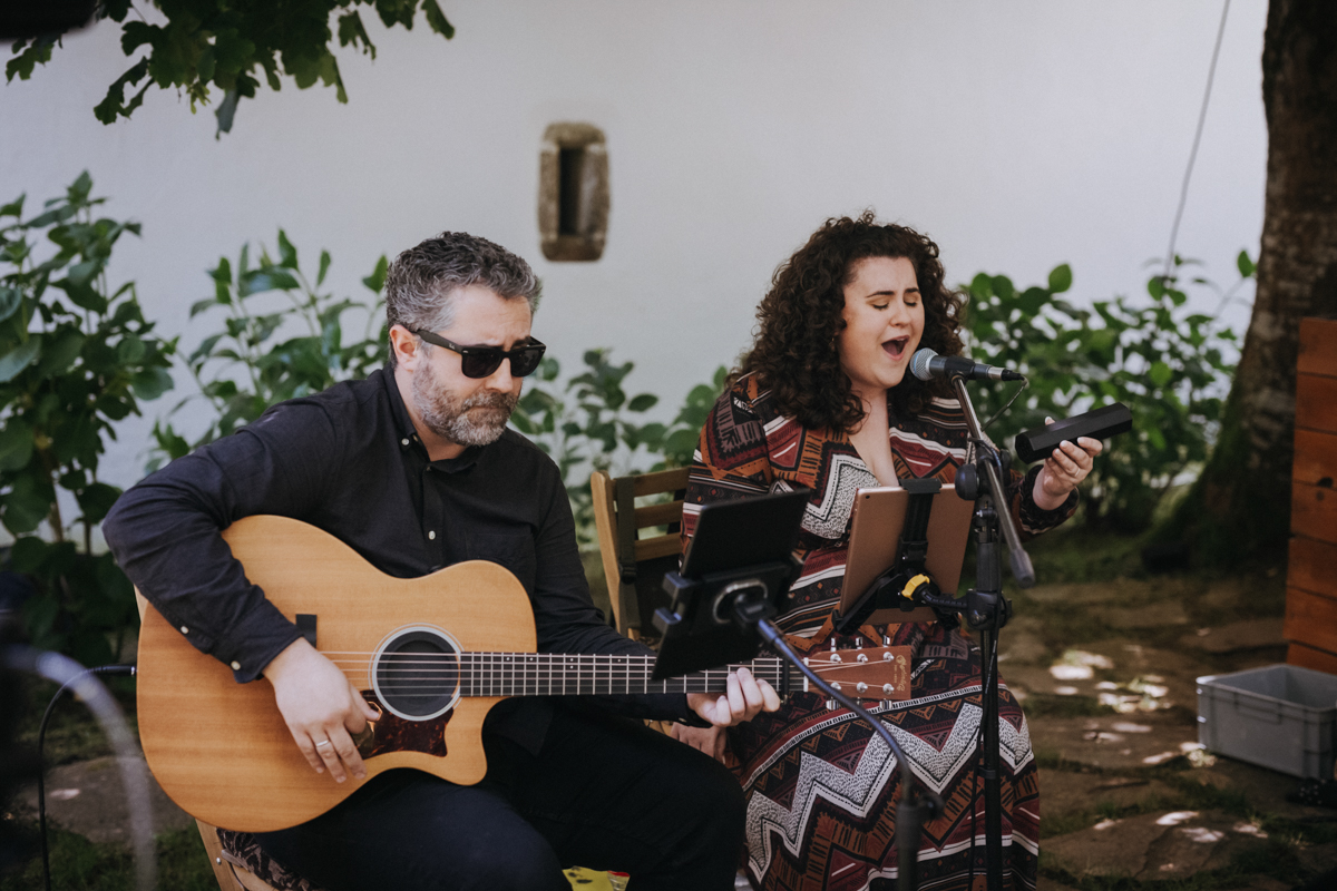 Noire et Blanche_429 Hombre toca guitarra acústica y mujer canta sentados en un jardín con pared blanca de fondo.
