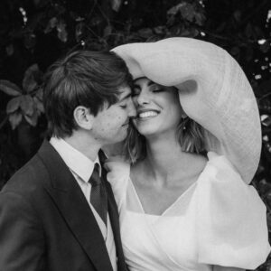 Pareja sonriente vestida de boda, ella con vestido blanco y sombrero grande, él con traje oscuro y corbata.