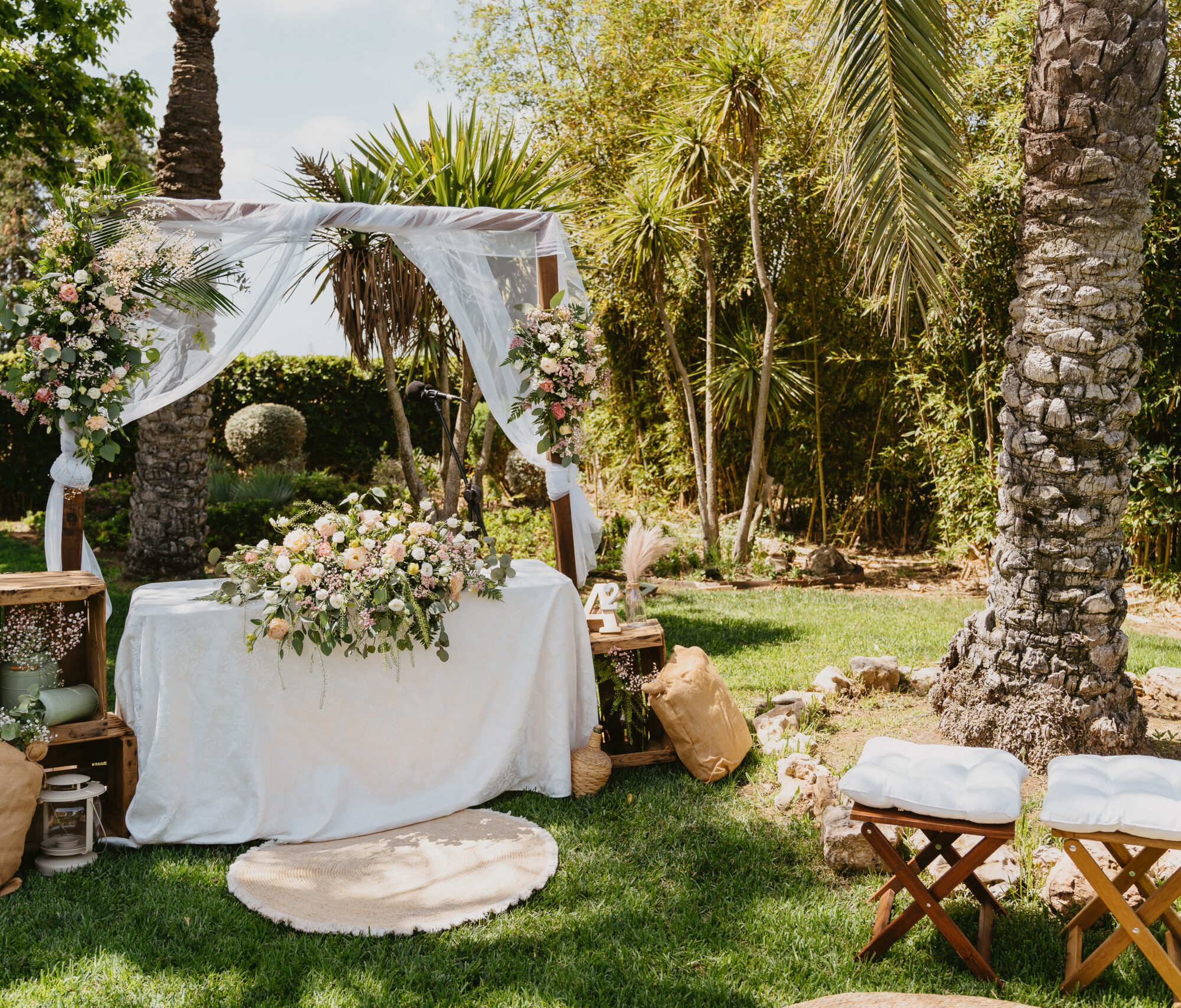 Team bride (6) Arco decorado con tela blanca y flores, mesa con mantel y arreglo floral en jardín soleado.