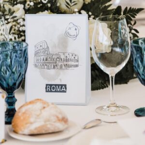 Mesa con copas, pan y tarjeta decorativa con dibujo del Coliseo y texto Roma.