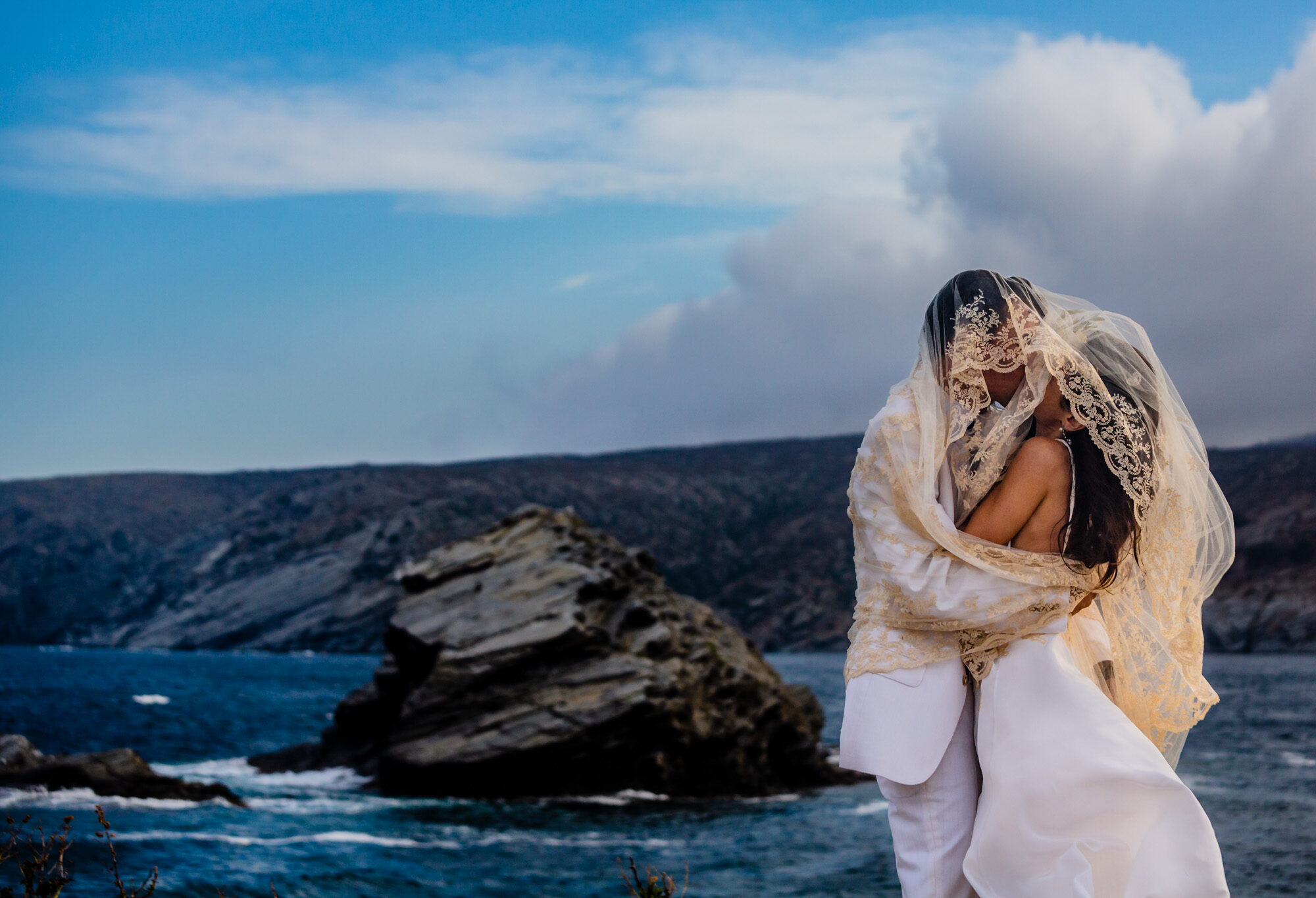 barcelona-wedding-photographer Pareja abrazada junto al mar con paisaje rocoso y cielo parcialmente nublado.