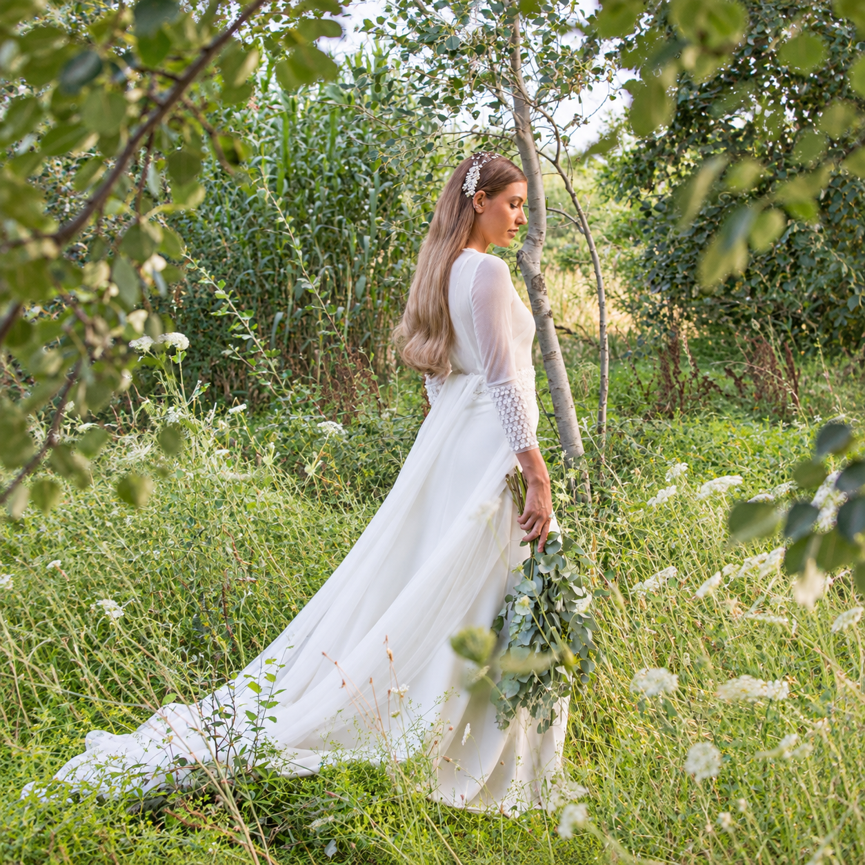 d63d2adc-b449-4229-a2d5-82013c077f97 Novia de perfil con vestido largo blanco y ramo, rodeada de vegetación en un campo al aire libre.