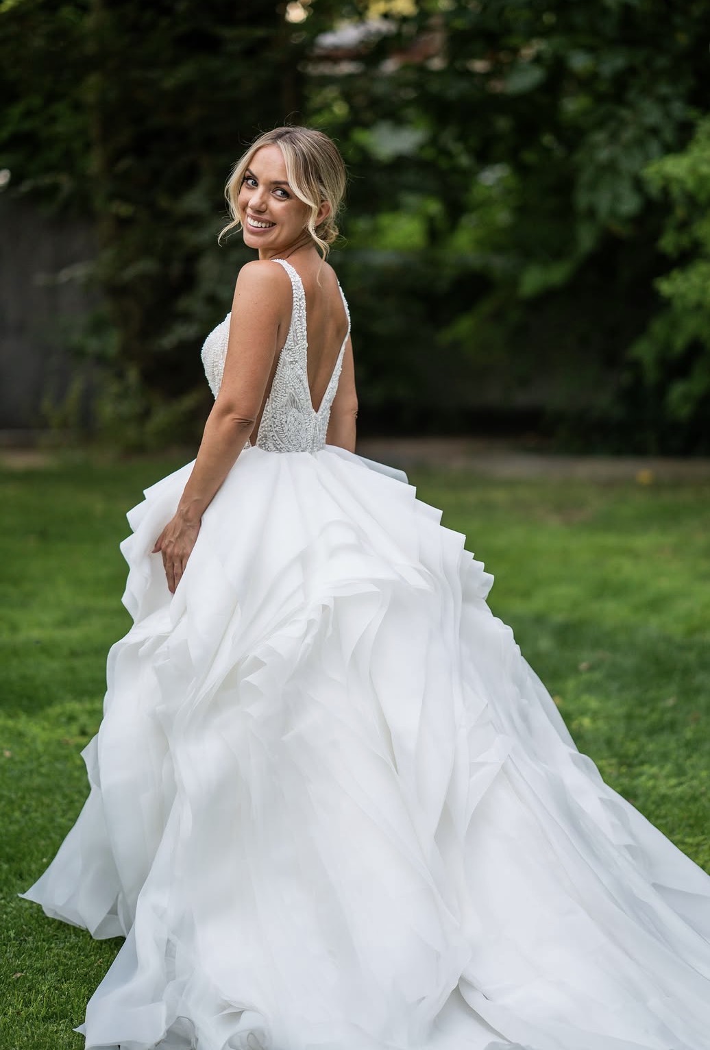 Screenshot Mujer sonriente con vestido de novia blanco de espalda descubierta en jardín verde.