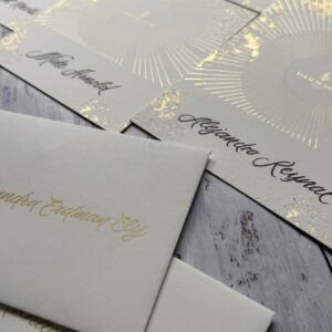 Sobres y tarjetas con caligrafía dorada sobre fondo claro y superficie de madera gris.